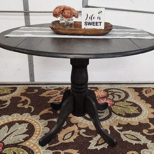 May include: A dark brown, round dining table with a pedestal base. The table has a decorative runner, a wooden bowl with flowers, and a sign that reads "Life is SWEET". The table is set on a patterned rug.