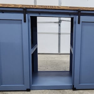 May include: A blue cabinet with sliding doors and a natural wood top. The cabinet has a rectangular opening in the center and black hardware. The doors slide on a black track. The cabinet is likely used for storage.