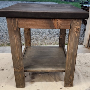 May include: A stained wooden side table with a dark top and a lower shelf. The table has four sturdy legs and a simple, rustic design. The wood grain is visible, adding to the natural aesthetic. The table is approximately 24 inches tall.