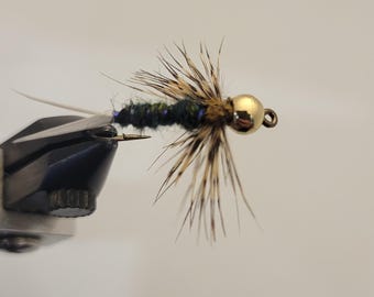 May include: A close-up of a handmade fishing fly. The fly features a gold bead head, a dark green body with blue accents, and brown and tan feather wings. The fly is attached to a silver hook.