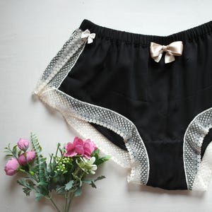 May include: Black underwear with white lace trim and small satin bows. The garment has a satin-like texture and a comfortable design. A sprig of pink and green flowers is in the lower left corner.