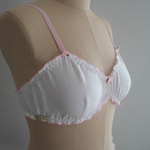 May include: White bra with pink trim and small rose embellishments. The bra has a gathered, ruffled design and a label that reads "BEUSKA".