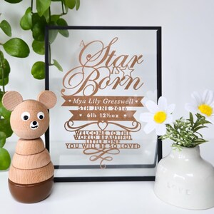 May include: A wooden bear stacking toy with a brown base and a framed birth announcement that reads "A Star is Born." The announcement includes the baby's name, birthdate, and weight (2.9 kg). A small white vase with daisies is also present.