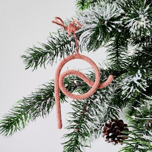 May include: A pink crocheted ornament shaped like a letter P hangs from a snowy artificial Christmas tree branch.