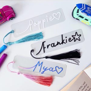 May include: Clear acrylic bookmarks with the names "Poppie", "Frankie", and "Mya" in cursive. Each bookmark has a tassel in blue, black, and pink. A heart and star are next to the names. Toy cars and markers are in the background.