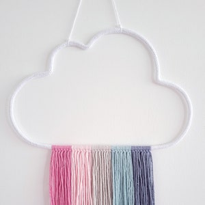 May include: A white metal cloud-shaped wall hanging with a rainbow of yarn strands hanging from the bottom. The yarn strands are pink, light pink, beige, light blue, and grey.