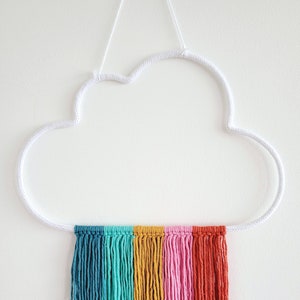 May include: A cloud-shaped wall hanging with a white frame and a rainbow of colourful fringe. The fringe includes shades of teal, green, yellow, pink, red, and orange. The wall decor is suspended by a white cord.