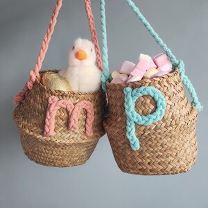 May include: Two woven seagrass baskets with braided handles, one pink and one teal. The basket on the left has a pink "m" and contains a plush duck and a gold egg. The basket on the right has a teal "p" and is filled with marshmallows.