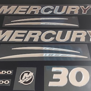 May include: A selection of chrome boat emblems and decals, featuring the brand name "MERCURY" in bold, stylized lettering. Also included are "VERADO" decals, a "300" emblem, and a logo with a stylized "M" and waves.