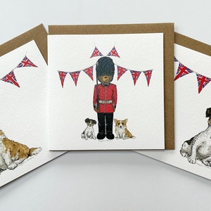 May include: Three greeting cards with a white background and brown envelopes. The cards feature illustrations of dogs and a British guard. The first card shows a corgi dog with a Union Jack bunting above. The second card shows a British guard with two dogs, a Jack Russell and a corgi, with a Union Jack bunting above. The third card shows a Jack Russell dog with a Union Jack bunting above.