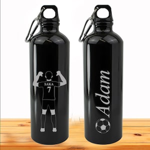 May include: Two black water bottles with a silver carabiner clip. One bottle has a silhouette of a football player with the text "SAKA 7" and the other bottle has the name "Adam" and a football graphic.