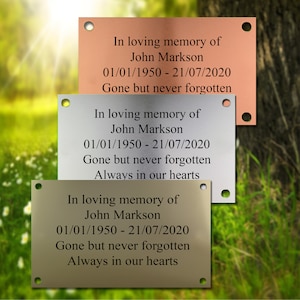 May include: Three metal memorial plaques with rounded corners and holes in the top corners. The plaques are in different colours: copper, silver, and gold. The text on the plaques reads: "In loving memory of John Markson 01/01/1950 - 21/07/2020 Gone but never forgotten Always in our hearts".
