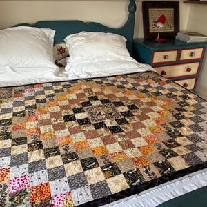May include: A handmade quilt with a patchwork design, featuring squares in various colors and patterns. The quilt is laid out on a bed with white pillows and a teal headboard. The quilt's colors include black, brown, orange, and floral prints.