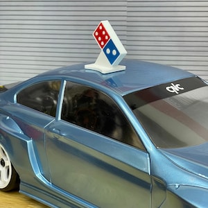 May include: A blue model car with a white and red Domino's Pizza logo on the roof. The car has a black windshield and a black sticker on the back that says "CNC".