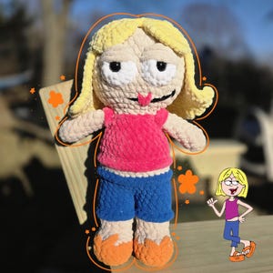 May include: A handmade crochet doll with yellow hair, a pink top, blue shorts, and orange shoes. The doll has a cartoonish design with large white eyes and a small tongue sticking out. The doll is standing on a wooden surface.