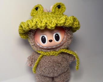 May include: A soft toy with a smiling face, wearing a lime green crocheted frog hat and matching scarf. The toy has a brown, furry body and small pink feet. The hat features two frog eye details.