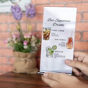 May include: A white sign with black text listing bar signature drinks: Cuba Libre, Gin Tonic, and Mojito. Each drink is illustrated with a glass of the cocktail.