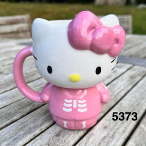 May include: A ceramic mug shaped like a white cat character with a large pink bow on its head. The mug features a yellow nose, black eyes, and whiskers. The base of the mug is pink and decorated with a white skeleton rib cage and bone print.