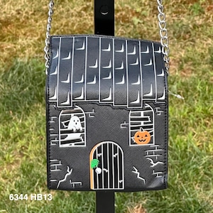 May include: A black crossbody bag shaped like a haunted house with a silver chain strap. The bag features a white ghost in a window, an orange pumpkin in another window, and a green hand on the door. The text 6344 HB13 is printed in white.