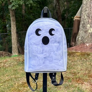 May include: A clear, transparent backpack featuring a black graphic of a ghost face with two crescent moon eyes and a circular mouth. The bag has black straps, a top handle, and is filled with white tissue paper, creating a ghostly appearance.