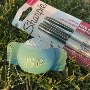 Op de afbeelding: Een golfbal sleutelhanger met de initialen "JSP" en twee zwarte Sharpie permanente markers. De Sharpie verpakking leest "FOR MARKING EVERYWHERE". De sleutelhanger is lichtblauw en groen, en de golfbal is wit.
