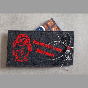 Könnte beinhalten: Eine graue Filz-Geschenkverpackung mit einem roten Zwerg, der ein Herz hält und den Text "kommt von Herzen". Die Tasche ist mit einem weißen und schwarzen Band und einem roten Marienkäfer verziert.
