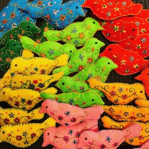 May include: A collection of felt bird ornaments in various colours, including red, green, yellow, blue, and pink. Each bird has embroidered details and a whimsical design.