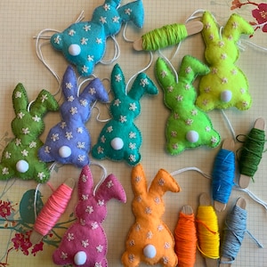 May include: Ten felt bunny decorations in a variety of colours, including green, blue, pink, and orange. Each bunny has a white pom-pom tail and is decorated with embroidered white flowers. The bunnies are hanging from white string and are paired with spools of embroidery floss in coordinating colours.