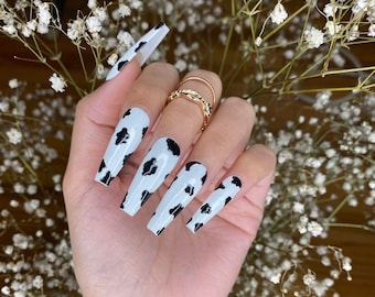May include: Cow print press-on nails with a white base and black spots. The nails are long and have a coffin shape.