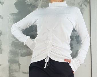 Puede incluir: Una camiseta blanca de manga larga con cuello alto y detalle de cordón, combinada con pantalones negros y zapatos de plataforma. El conjunto se completa con zapatillas de plataforma blancas y negras.