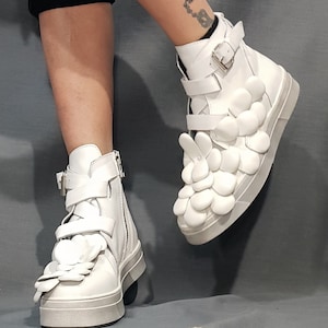 May include: White high-top sneakers with a unique design featuring multiple white flower-shaped embellishments on the upper and sides. The shoes have a white sole and a zipper on the side.