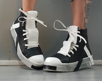 Op de afbeelding: Zwart-witte high-top sneakers met een uniek ontwerp met een witte band over de bovenkant van de schoen en een witte streep aan de zijkant. De schoenen hebben een dikke witte zool.