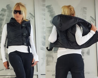 May include: A black puffer vest with a hood and buckle closures. The vest is cropped and has a quilted design. The model is wearing black jeans and platform sneakers.