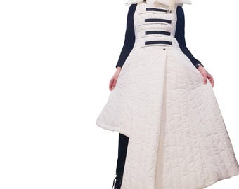 May include: A long, white, quilted sleeveless coat featuring a high collar, an asymmetrical hemline, and four horizontal black straps across the chest. The garment is worn over a black long-sleeved top, black pants, and black platform boots.