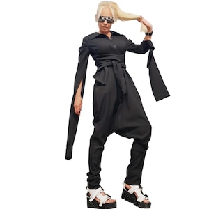 May include: A black jumpsuit featuring a collared neckline, a front zipper, and long sleeves with split cuffs. The garment includes a wrap-around waist tie, a drop-crotch design, and tapered legs. The outfit is paired with white platform sandals.