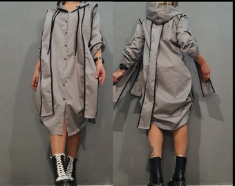 Puede incluir: Un vestido camisa gris con capucha y ribetes negros y cierre de botones. El vestido tiene un corte holgado y se muestra en una modelo con botas de plataforma negras.