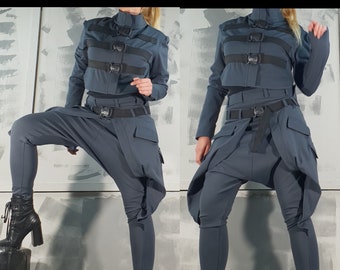 May include: A two-piece outfit in a dark gray color. The top is a cropped jacket with a high collar and multiple black straps with buckles. The pants are loose-fitting with a wide leg and a black belt. The model is wearing black heeled boots.