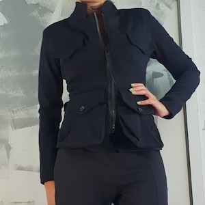 May include: A black, fitted, zip-up jacket with a high collar and multiple pockets. The jacket is paired with black trousers that have a slit at the bottom of each leg.