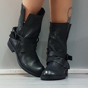 May include: Black leather ankle boots with a low heel. The boots feature a wrap-around strap with a buckle and a folded-over design at the top. The boots are worn by a person, and the background is a solid gray.