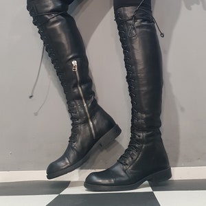 May include: Black leather over-the-knee boots with a lace-up design and a side zipper. The boots have a flat sole and a sleek, modern aesthetic. The boots are shown against a gray and black checkered background.