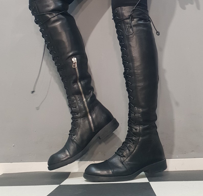May include: Black leather over-the-knee boots with a lace-up design and a side zipper. The boots have a flat sole and a sleek, modern aesthetic. The boots are shown against a gray and black checkered background.