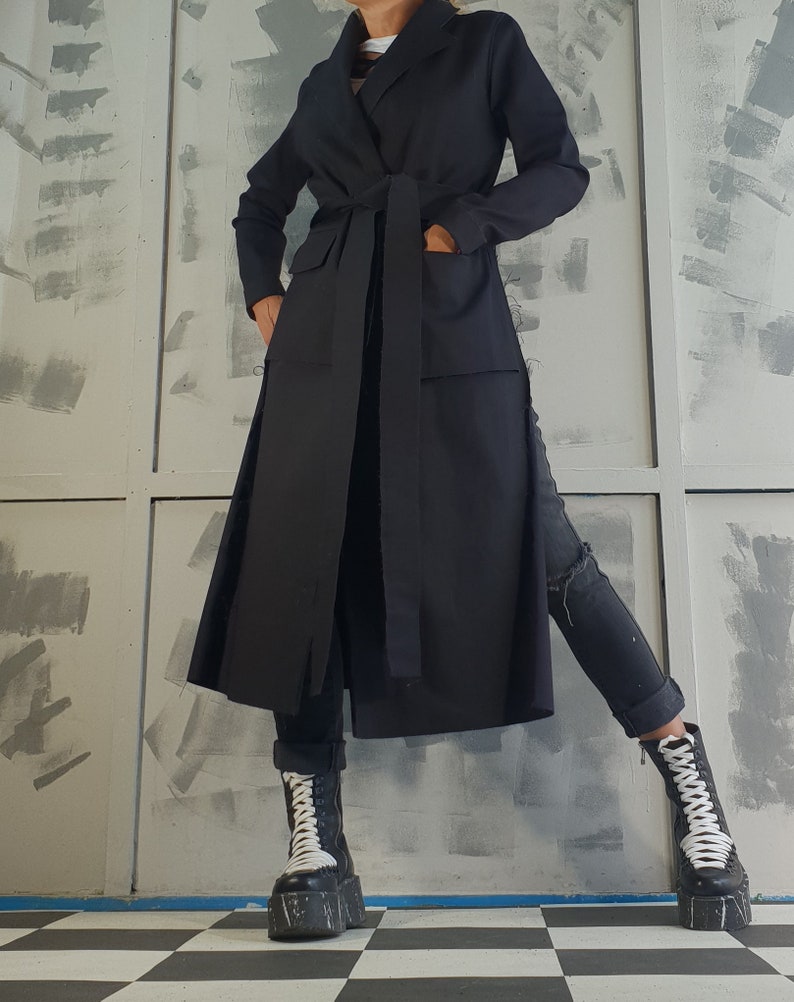 May include: A long black coat with a tie belt and pockets. The coat is worn over black jeans and black and white platform boots.