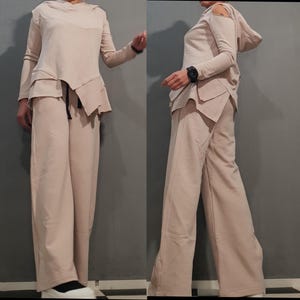 Può includere: Un completo beige a due pezzi con un top a maniche lunghe con un design asimmetrico a strati e un cappuccio. I pantaloni a gamba larga coordinati hanno una vita alta. L'outfit è realizzato in un tessuto morbido e fluido.
