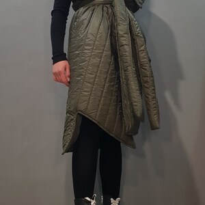 May include: A long, quilted, green vest with a large collar and a tie belt. The vest is worn over a black long-sleeved shirt and black leggings. The person is wearing black platform boots with white laces.