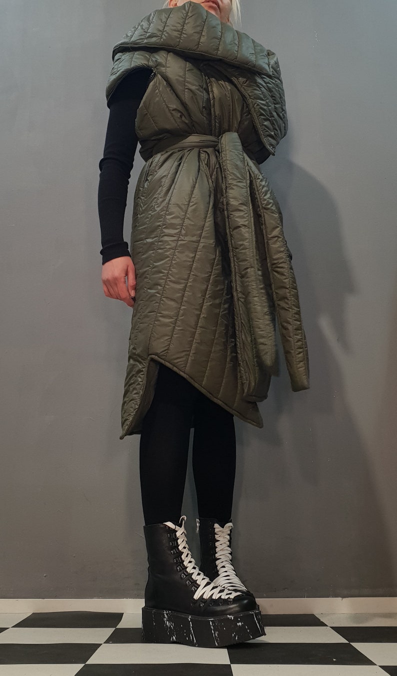 May include: A long, quilted, green vest with a large collar and a tie belt. The vest is worn over a black long-sleeved shirt and black leggings. The person is wearing black platform boots with white laces.