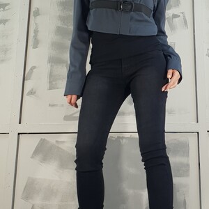 May include: A woman wearing a gray cropped jacket with black straps and buckles, black jeans, and black heeled boots with laces.