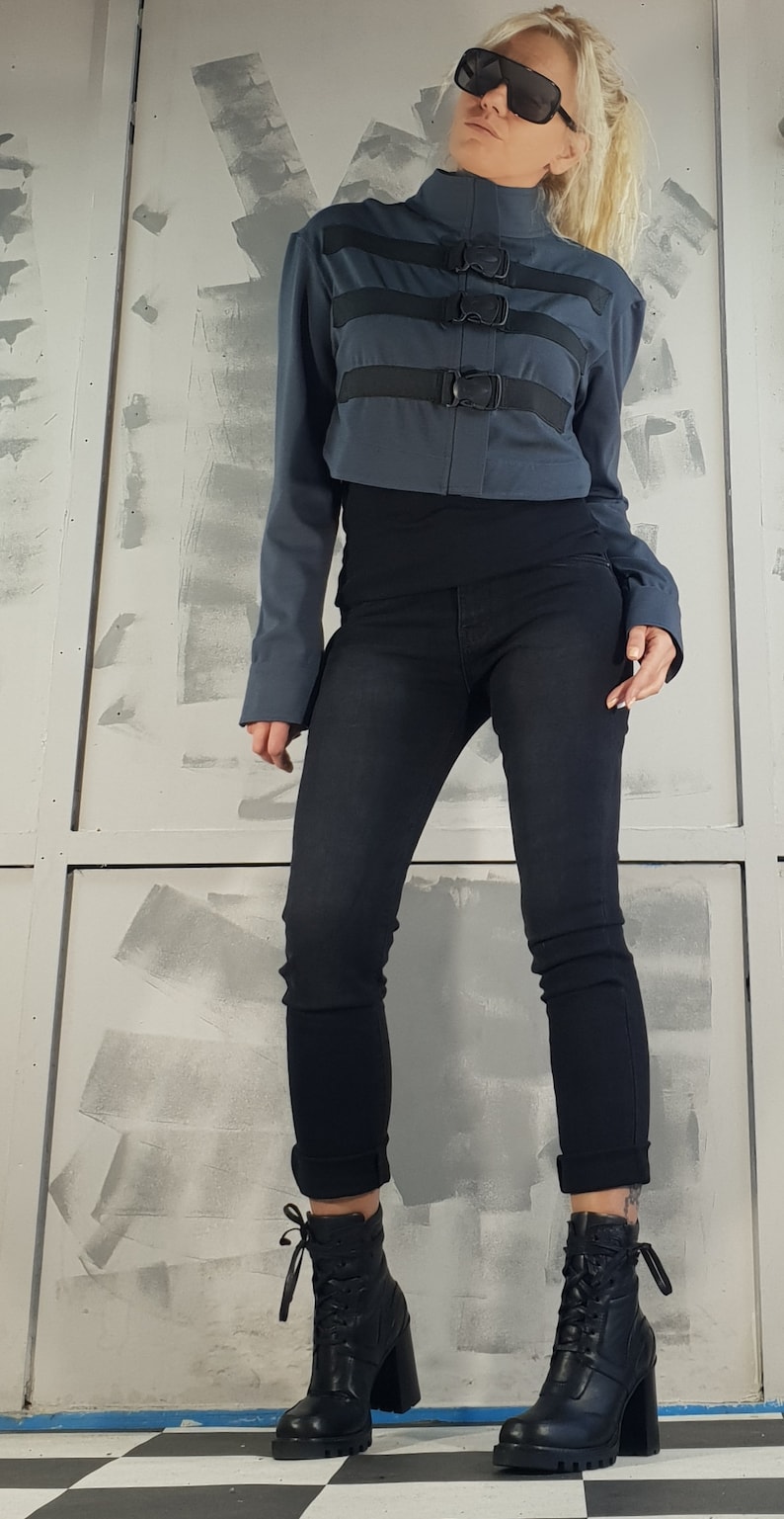 May include: A woman wearing a gray cropped jacket with black straps and buckles, black jeans, and black heeled boots with laces.