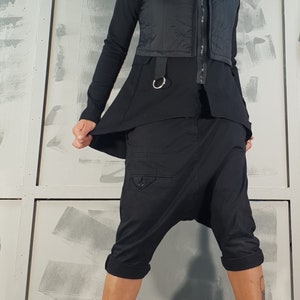 May include: A woman wearing a black vest, black pants, and black boots. The vest has a zipper and a belt with a silver ring. The pants are loose-fitting and have a cargo pocket on the left leg. The boots are lace-up and have a thick platform.
