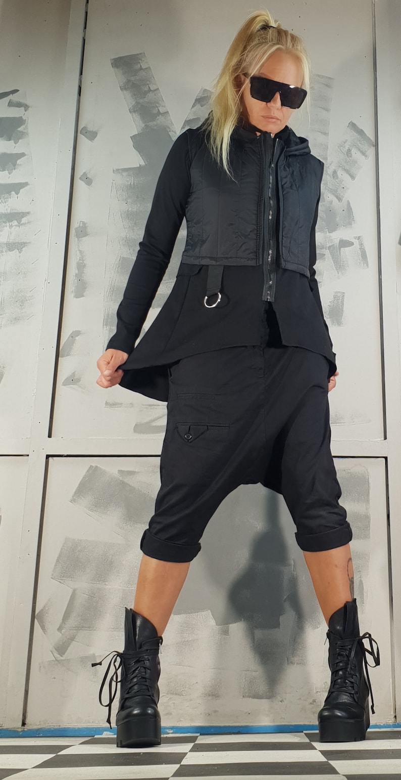 May include: A woman wearing a black vest, black pants, and black boots. The vest has a zipper and a belt with a silver ring. The pants are loose-fitting and have a cargo pocket on the left leg. The boots are lace-up and have a thick platform.