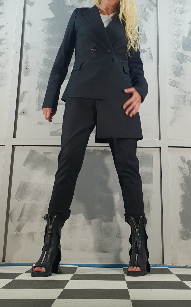 May include: A black blazer with a double-breasted closure and a matching pair of pants. The blazer has a longer back hemline than the front. The person is wearing black heeled boots with zippers.